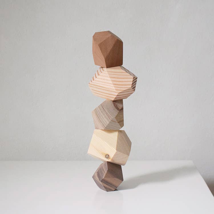 Five faceted wooden balance stones stacked into a leaning tower, each cut from a different wood, from dark walnut to pale pine, on a white surface