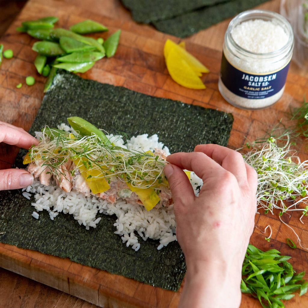 Hands rolling sushi on a wooden board beside an open jar of Jacobsen Garlic Salt