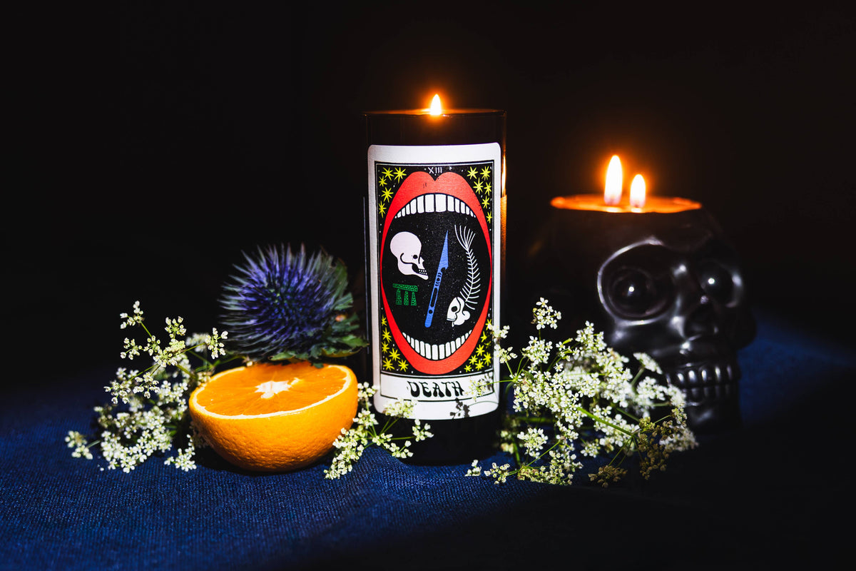 Tarot Candle, Death, burning beside a small skull, dried flowers and an orange half on a dark blue cloth