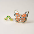 Two enamel pins, a striped green caterpillar and an orange and black monarch butterfly with white spotted edges, on a cream background