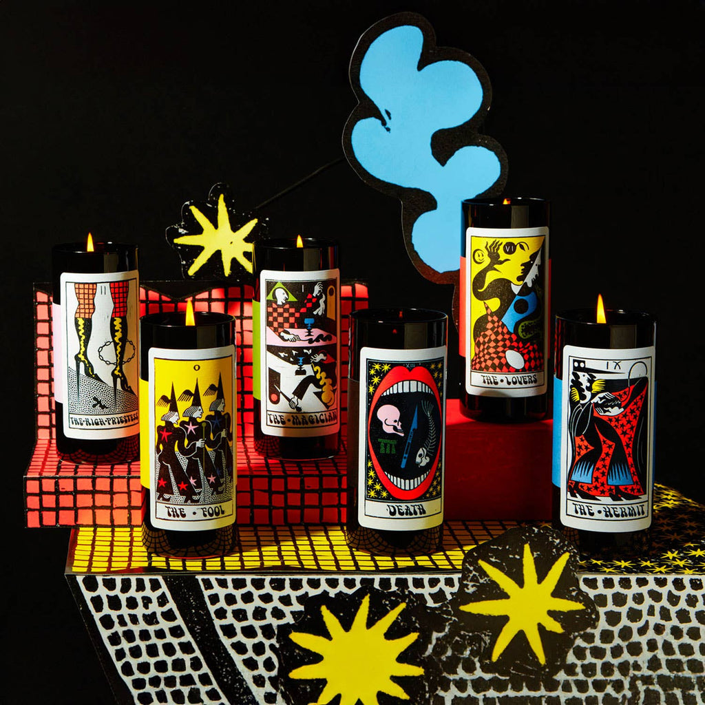 Several candles from the Tarot Candle collection lined up on a table with graphic paper props
