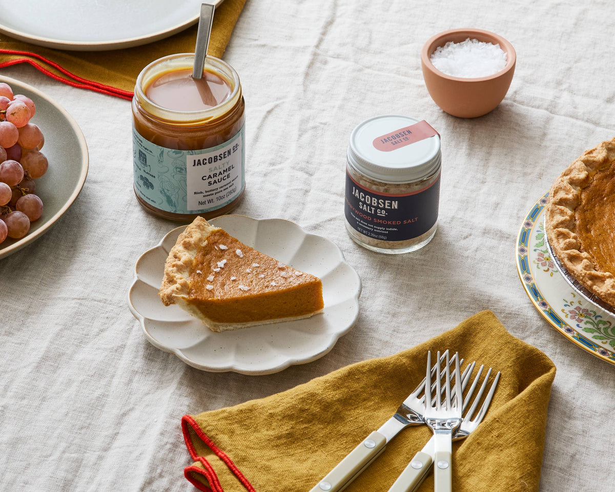 Table set for dessert with a jar of Jacobsen Cherrywood Smoked Salt, a small bowl of flake salt and slices of pie