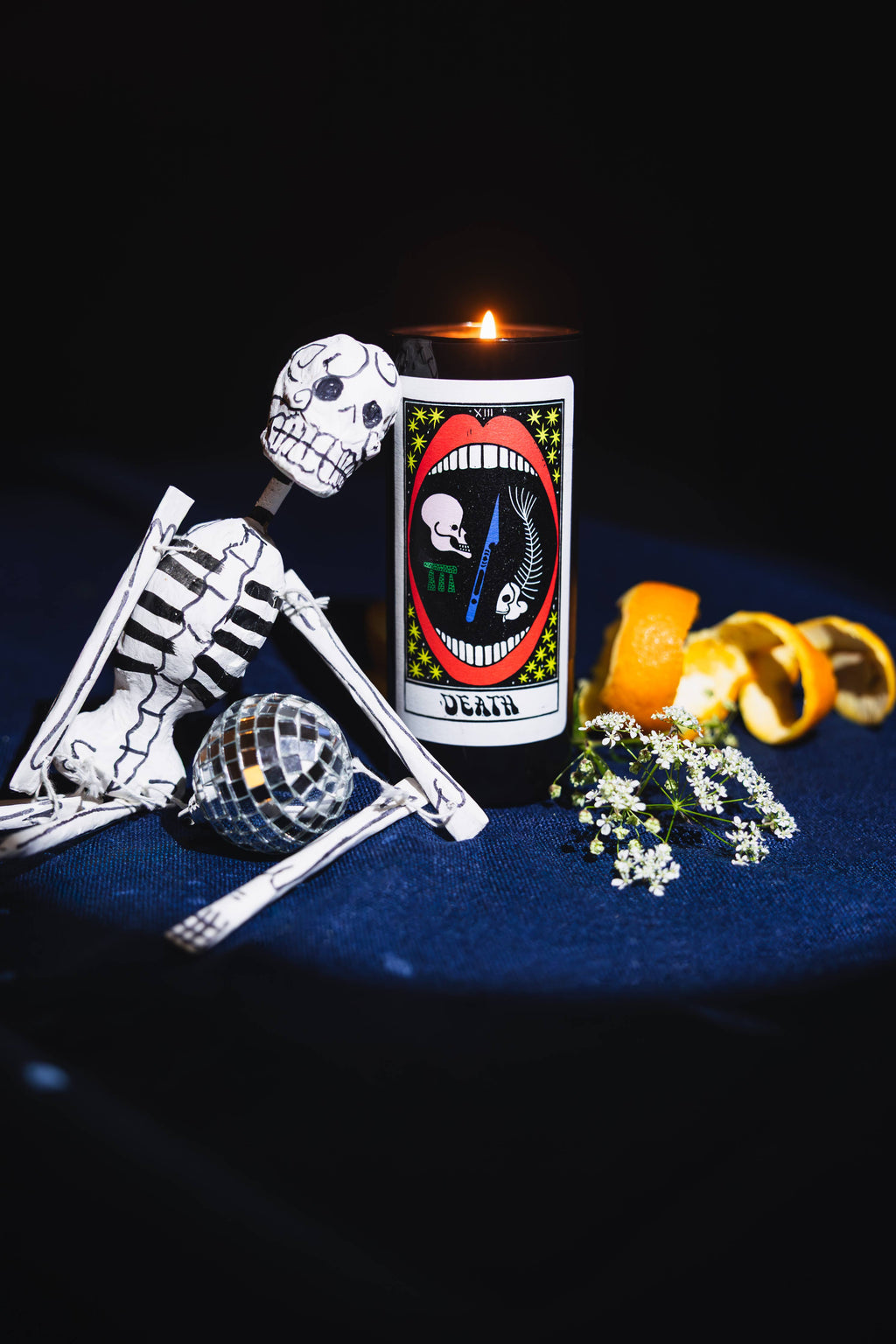 Tarot Candle, Death, lit on a dark blue ground beside two small skeleton figures and curls of orange peel