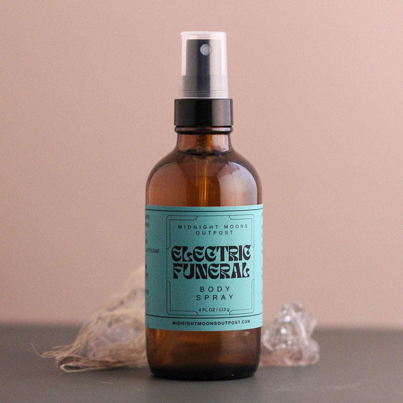 Electric Funeral body spray by Midnight Moons Outpost, amber glass with a turquoise label, beside clear quartz