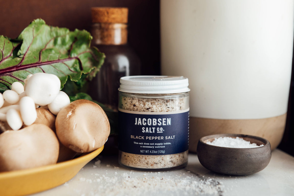 Jar of Jacobsen Black Pepper Salt on a kitchen counter beside mushrooms, beet greens and a small bowl of flake salt