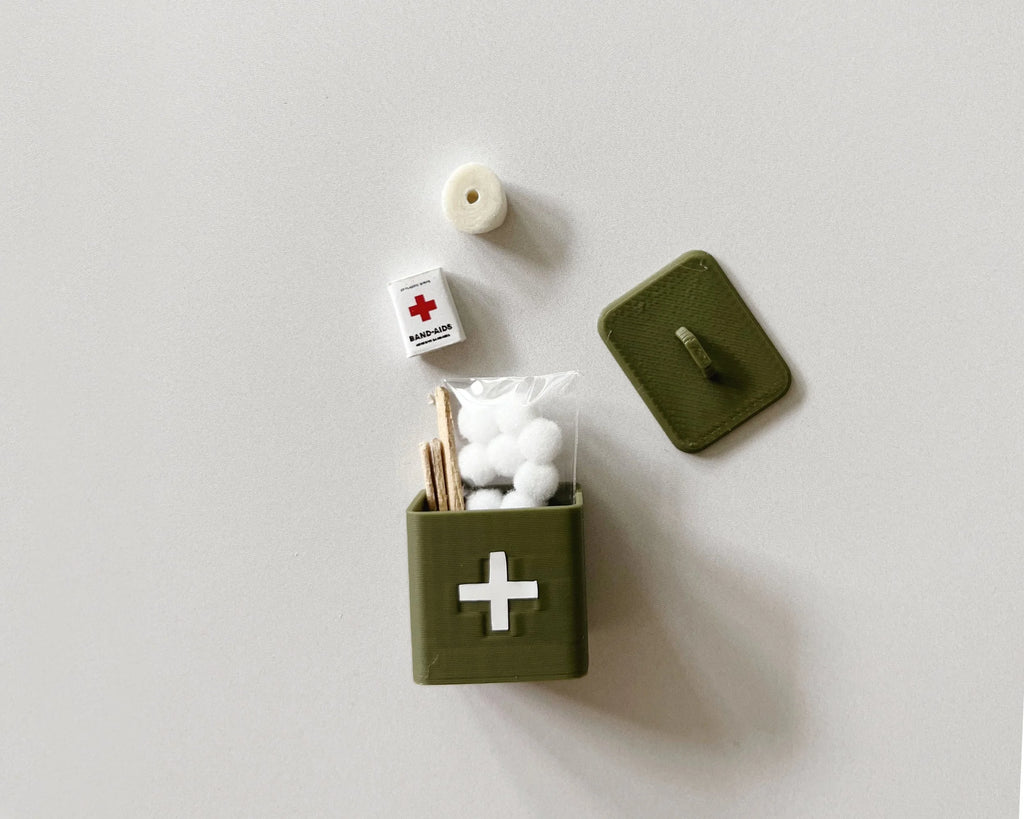 Olive miniature first aid box with lid off beside its tiny bandage box, cotton balls, tape roll and tongue depressors