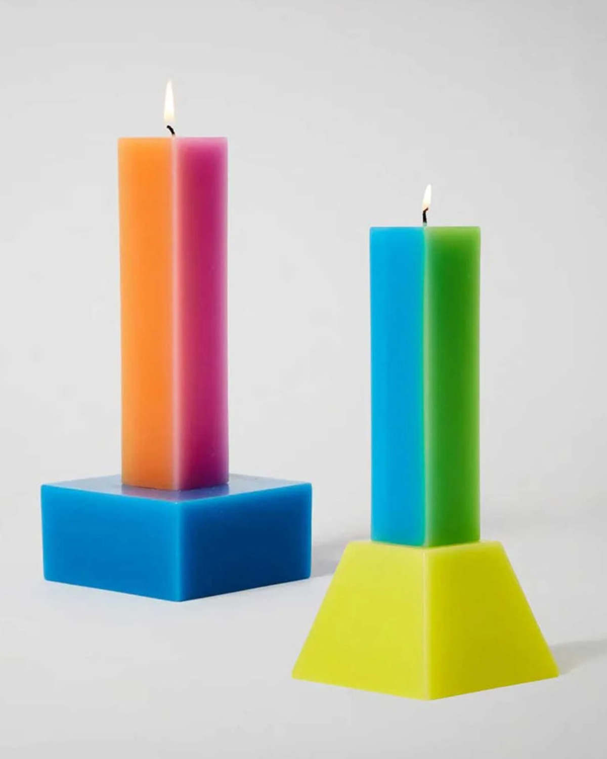 Both Yinka Ilori Happiness pillar candles lit side by side, orange and pink on a blue base next to blue and green on a yellow tapered base