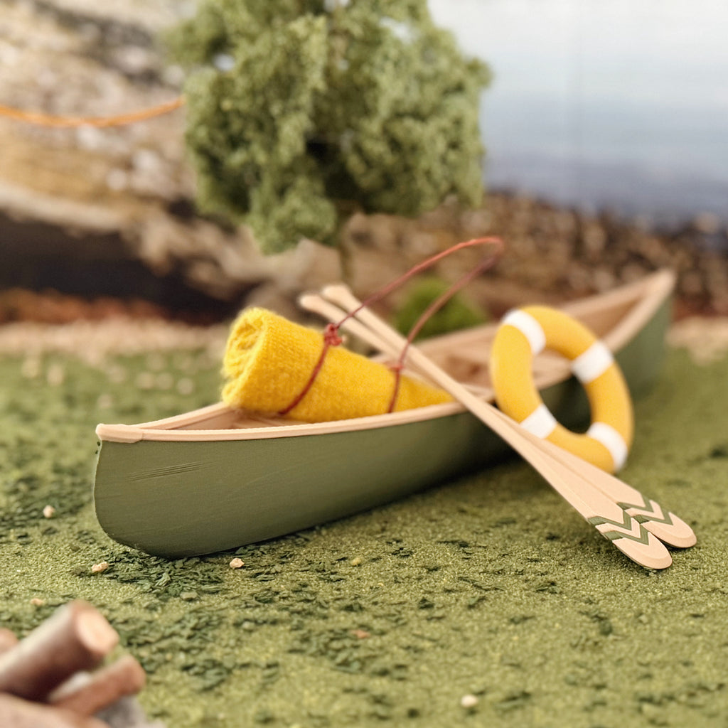 Green miniature canoe pulled up on a mossy bank with crossed oars, a rolled mustard blanket and a life ring inside