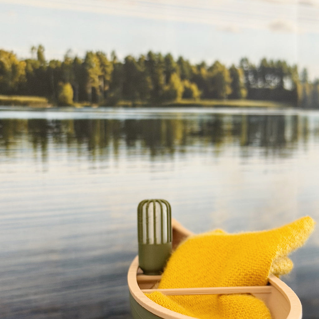 Stern of the miniature canoe in front of a lake and pine treeline, with a tiny thermos and a mustard blanket aboard