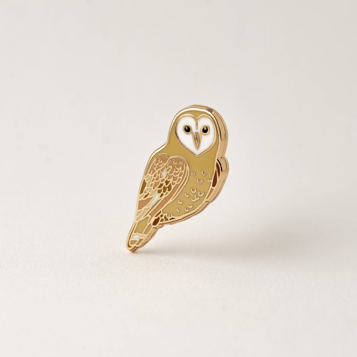 Barn owl hard enamel pin with a white heart shaped face and tan and caramel feathers outlined in gold, on a cream background