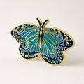 Blue morpho butterfly enamel pin with teal, blue and navy wings dotted in pale blue, outlined in gold, on a cream background
