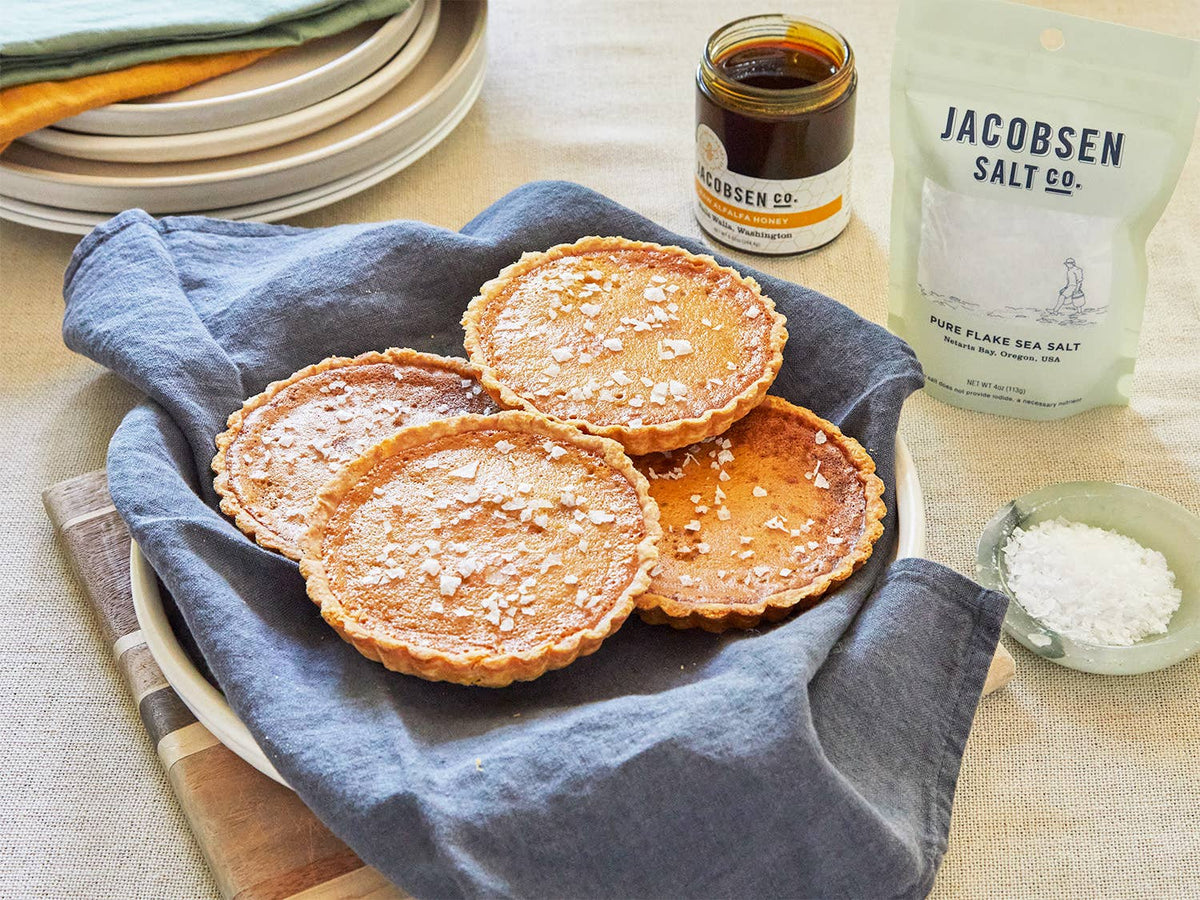 Jacobsen Raw Alfalfa Honey and a bag of flake sea salt beside small custard tarts in a blue napkin