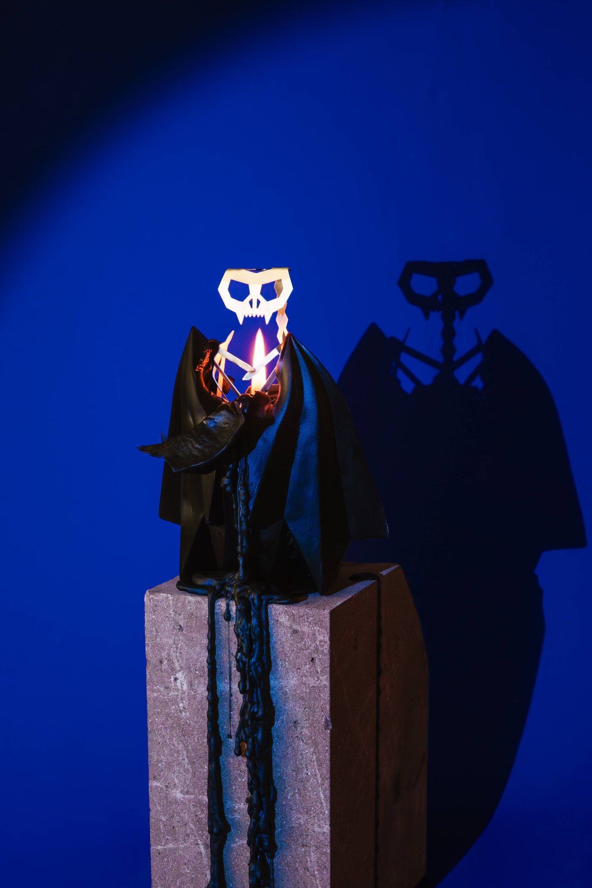 Blaka bat candle burning on a plinth in deep blue light, its shadow thrown on the wall behind