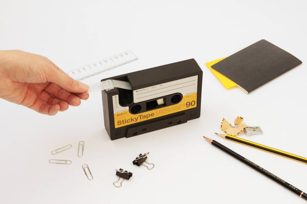 Cassette tape dispenser on a desk, a black and yellow cassette shape dispensing clear tape beside paper clips and a pencil