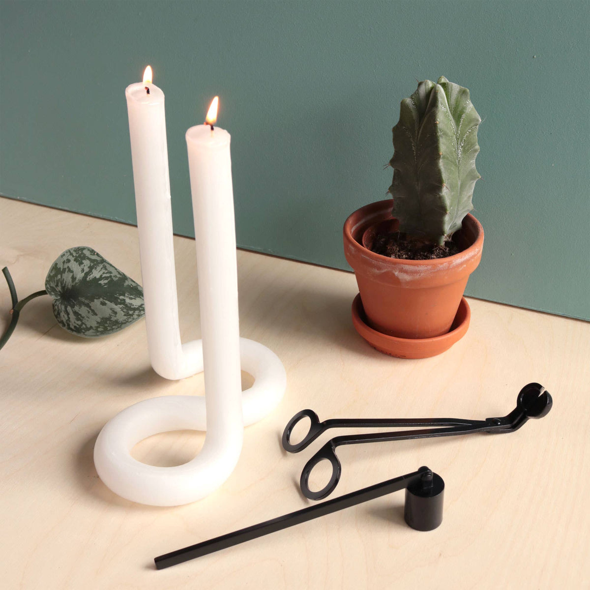 The snuffer and trimmer on a table beside two white tapers in a curved holder and a potted cactus