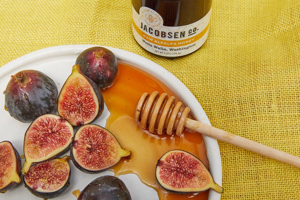 Jar of Jacobsen Raw Alfalfa Honey behind a plate of halved figs with a honey dipper on a yellow cloth
