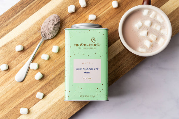 Mint green Moonstruck Minty hot cocoa tin on a wooden board beside a mug of cocoa with marshmallows and a spoonful of cocoa mix