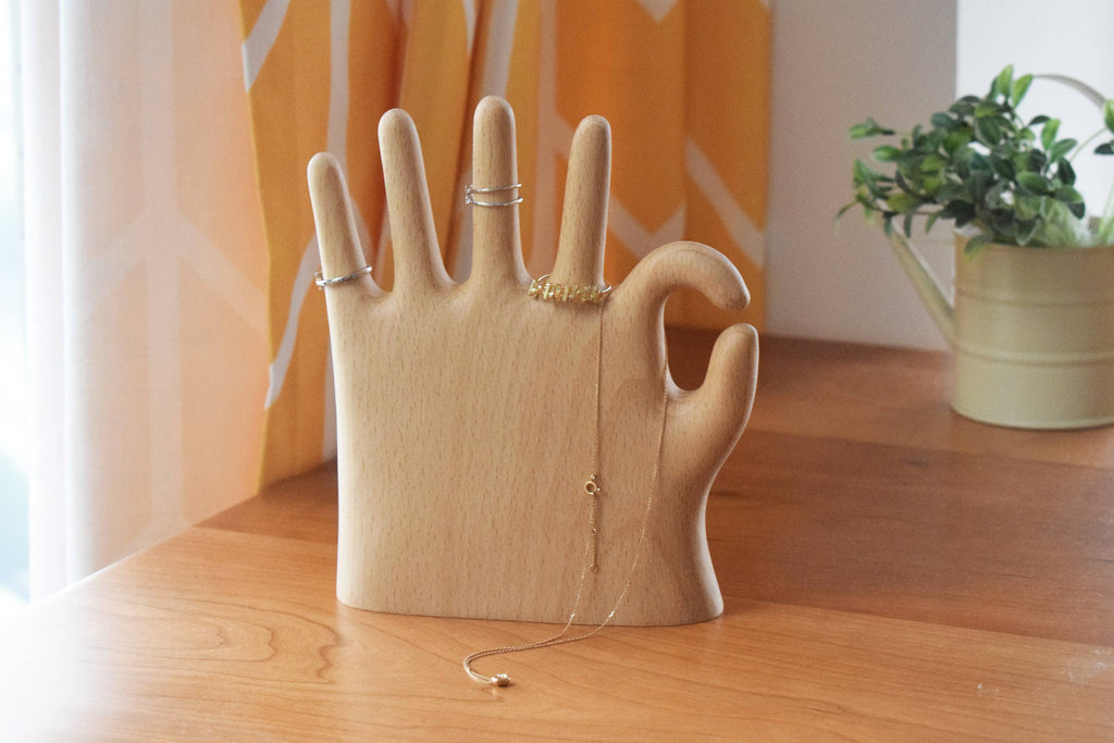 OK hand jewelry stand on a walnut desk with rings on the fingers and a necklace draped over the top, orange curtain behind