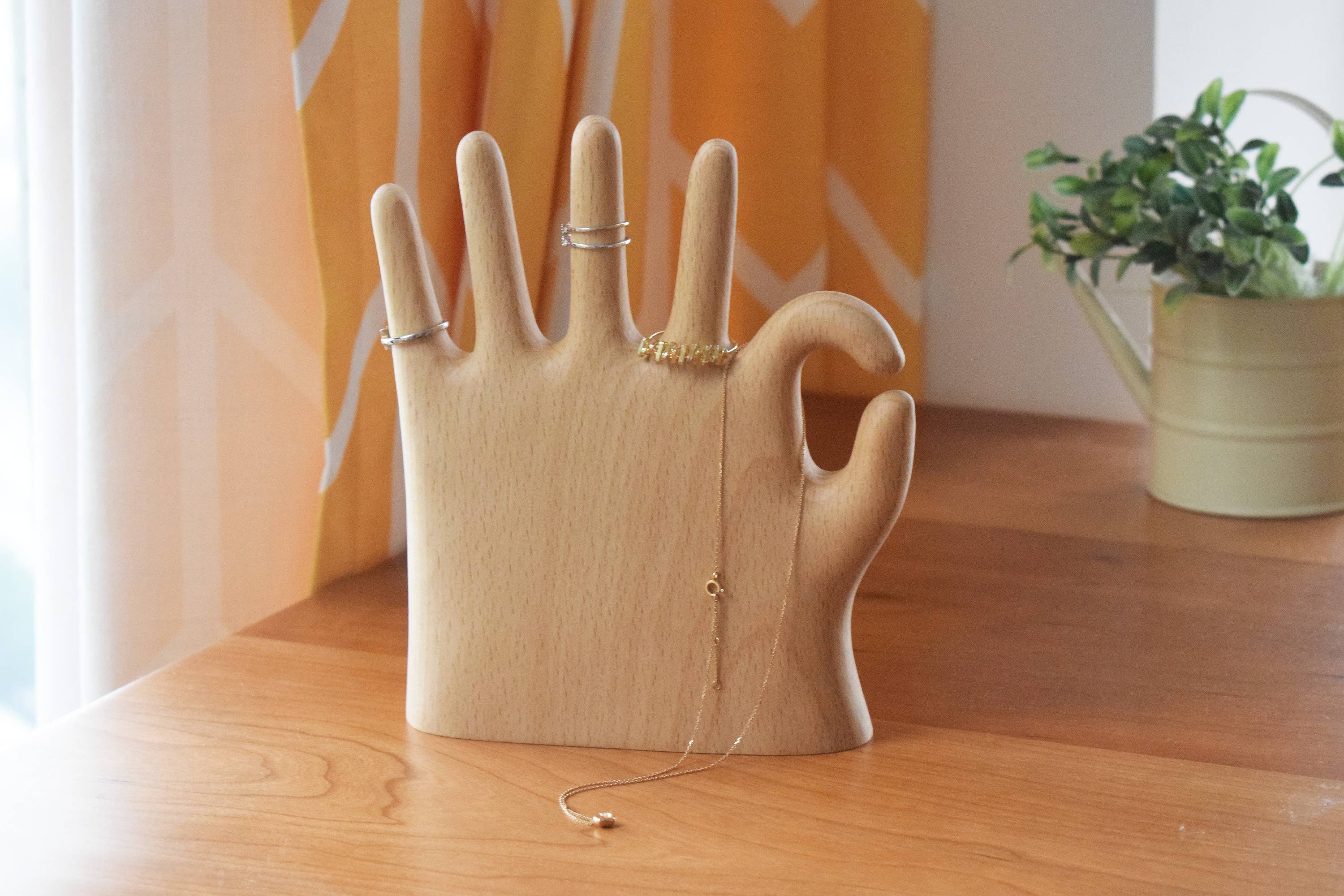 OK hand jewelry stand on a walnut desk with rings on the fingers and a necklace draped over the top, orange curtain behind
