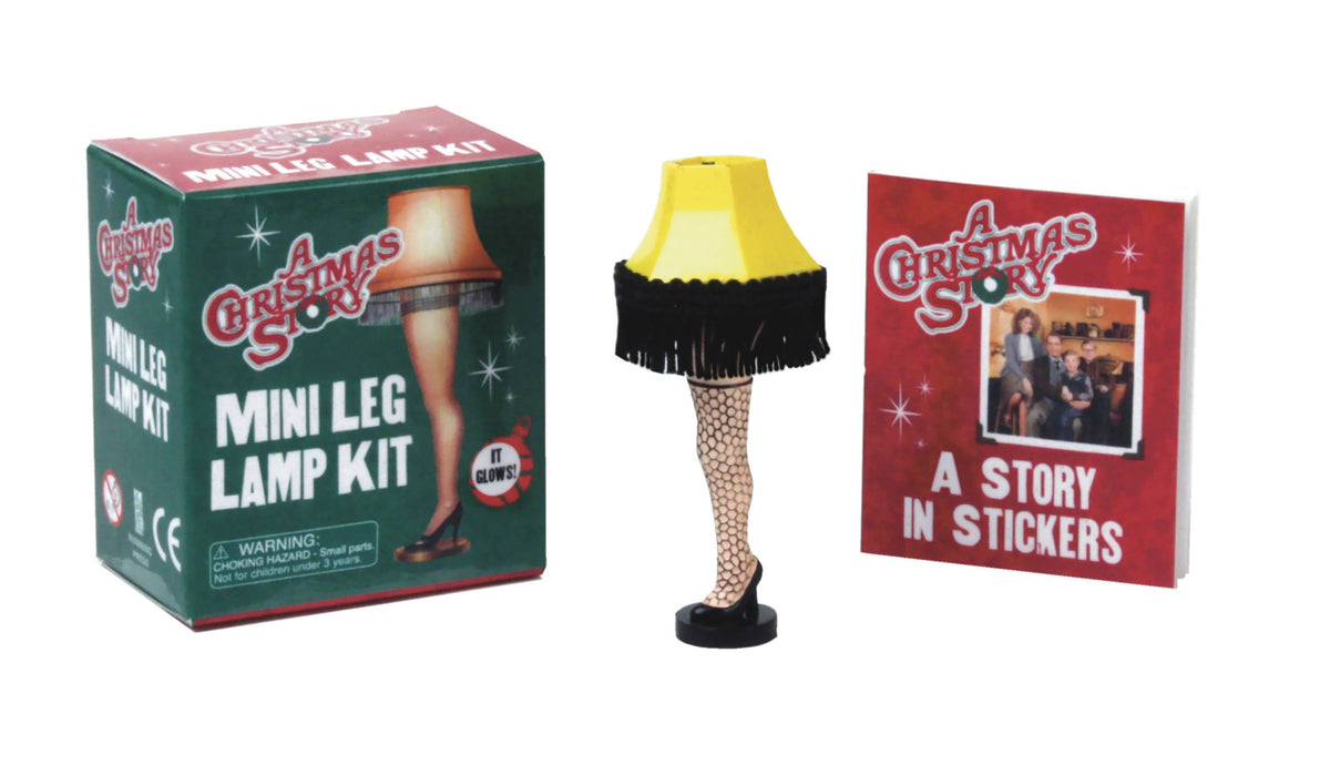 A Christmas Story mini leg lamp: a tiny lamp shaped like a leg in a fishnet stocking with a fringed yellow shade, between its green box and a sticker book