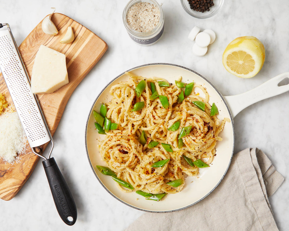 Overhead of a pan of pasta with a jar of black pepper salt, peppercorns and half a lemon on a marble counter