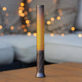 Slim grey perforated metal candle light glowing warm amber on a wooden table, string lights blurred behind it