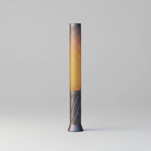 Slim grey perforated metal candle light glowing warm amber on a wooden table, string lights blurred behind it
