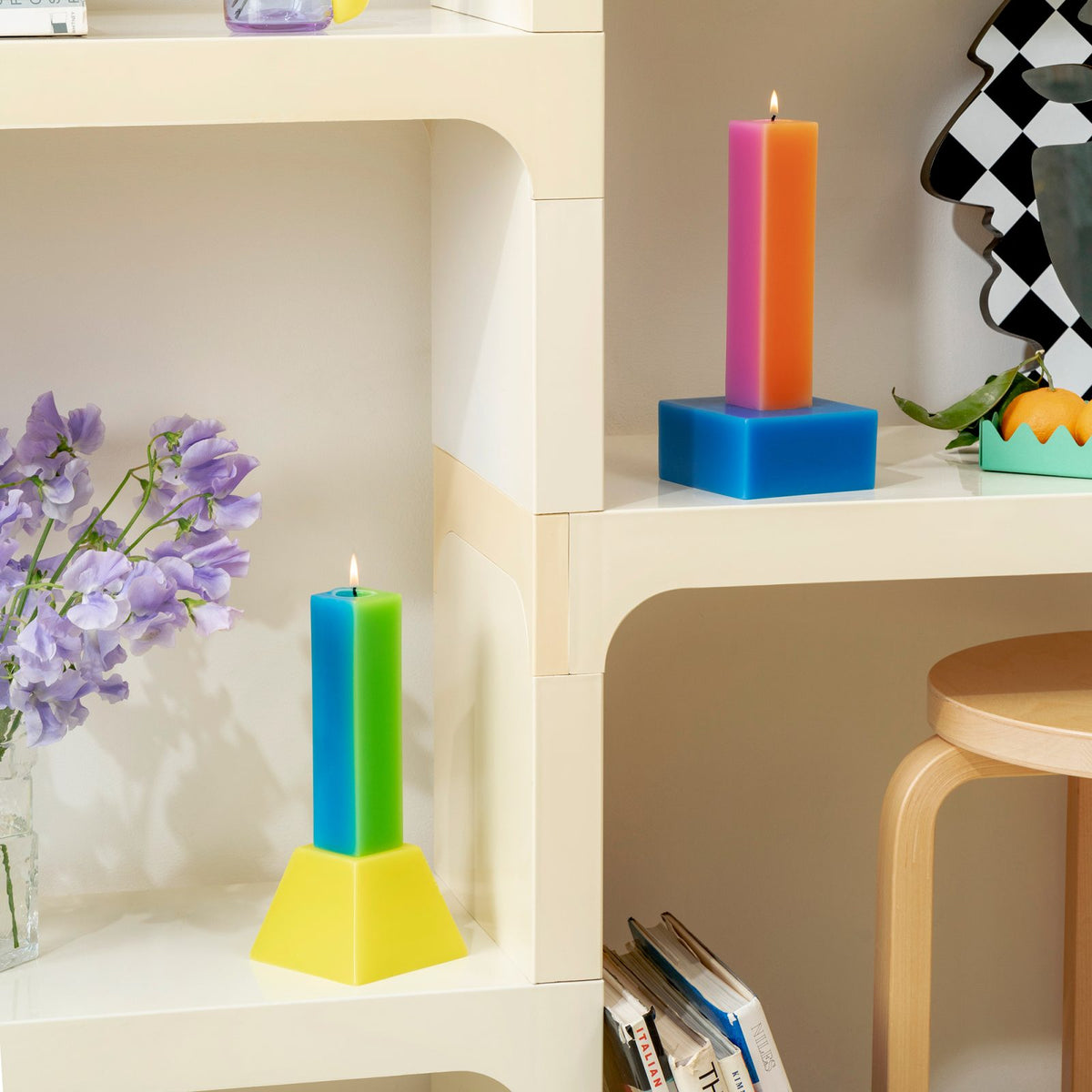 Both Happiness candles lit on white modular shelving beside lilac flowers and a bentwood stool