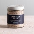 Jar of Jacobsen Black Pepper Salt, 4.23 oz, with a navy label and white lid on a pale stone table