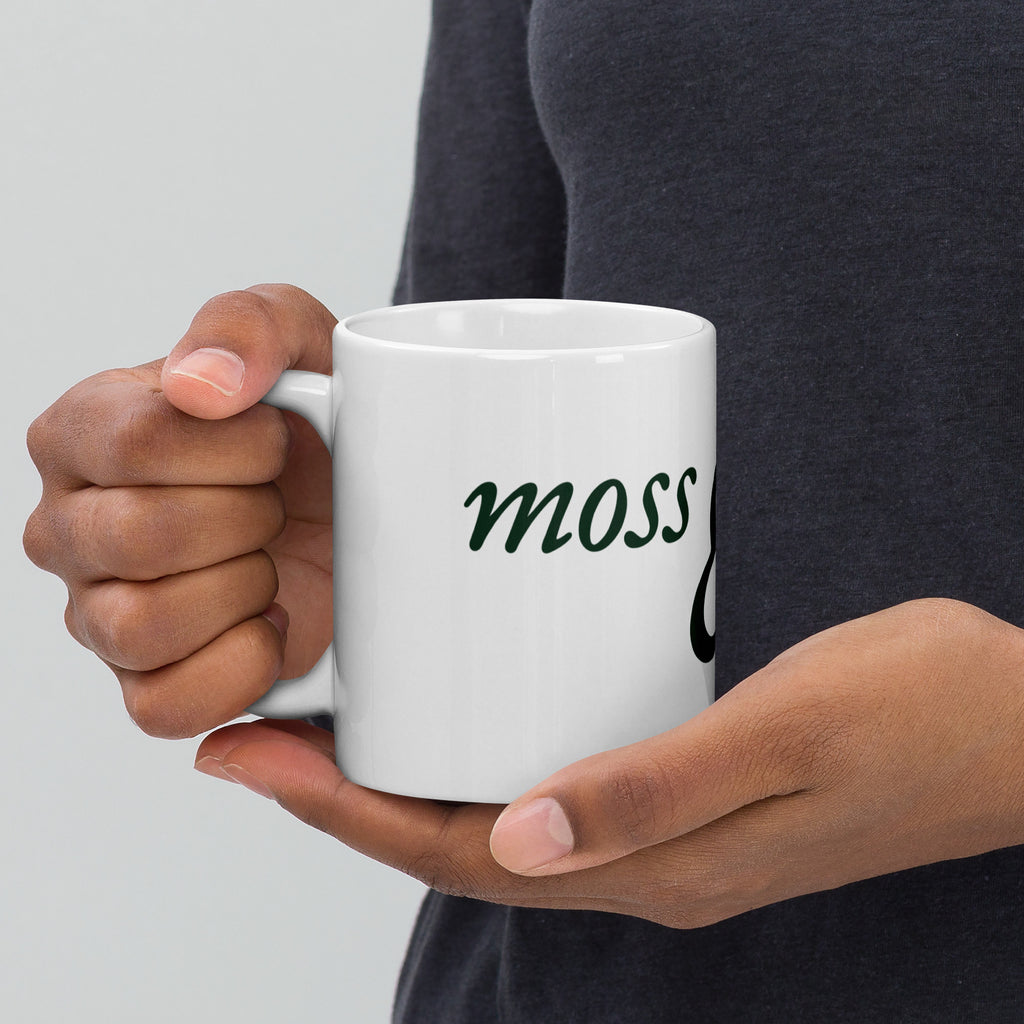Hands holding a white glossy mug printed with the Moss and Fog wordmark in green script