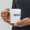Hands holding a white glossy mug printed with the Moss and Fog wordmark in green script